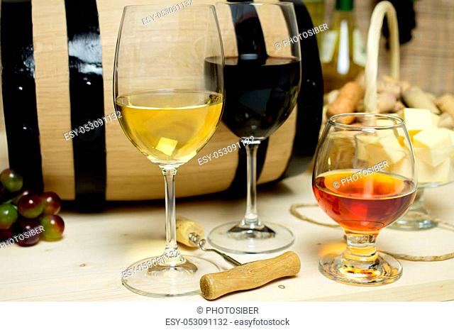 Red and white wine in glasses, brandy. On the background of a barrel and wine corks basket, bowl with cheese and a bottle of wine, a corkscrew