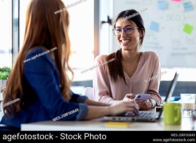 Smiling businesswomen using smartphone, talking with her colleague