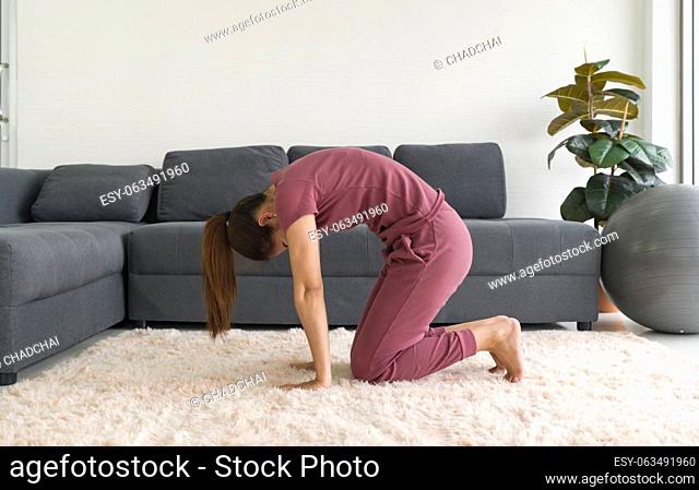 Asian housewife doing Cat Pose exercise during home workout. Female practicing yoga in Marjaryasana Pose on carpet in living room