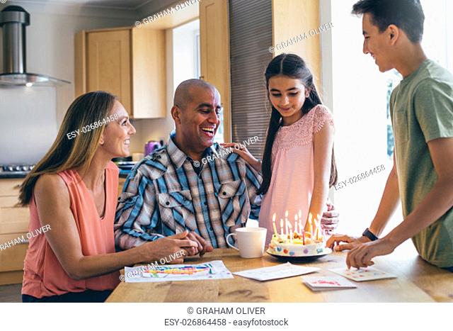 Family of four celebrating the fathers birthday with a cake that the children made. He is laughing as they sing before he blows out the candles