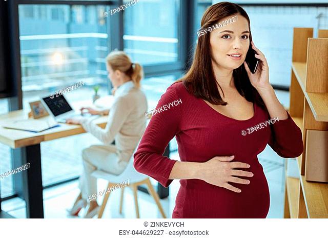 Expecting baby. Pretty female person standing on the foreground and putting right hand on the belly while calling to her doctor