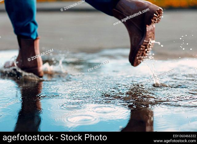 Woman wearing rain rubber boots walking running and jumping into puddle with water splash and drops in autumn rain season