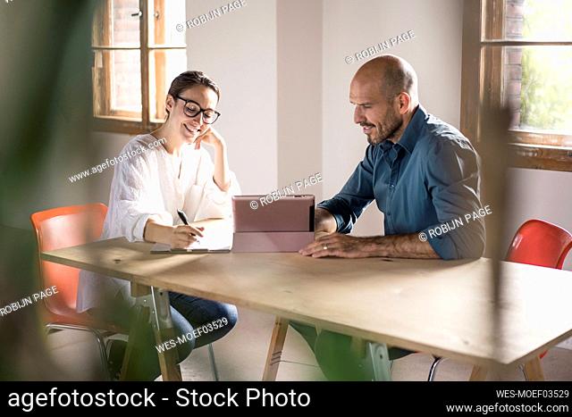 Businesswoman writing in notepad while sitting by man using digital tablet at office