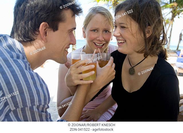 Ein junger Mann und zwei junge Frauen prosten sich mit ihren Cocktails am Sandstrand zu Model M.: Lara, Model r.: Marlene
