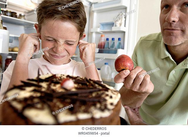 Boy standing in front of a cake with his fingers in his ears and his father holding an apple
