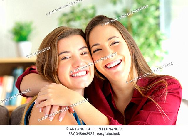 Front view portrait of a two happy friends or sisters posing smiling and looking at you with a homey background