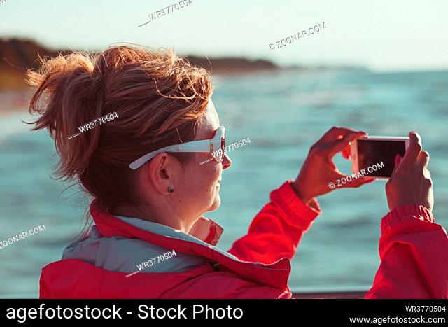 Young woman taking a photos using phone, looking at screen, standing outdoors, she is backlighted by sunlight with plain sky and sea in the background