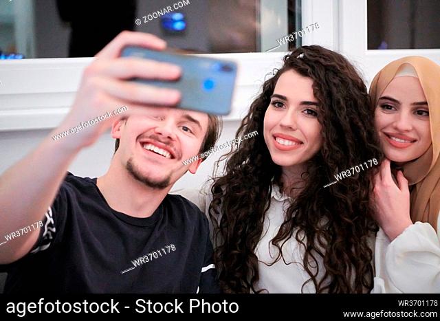 group of young muslim people one woman in fashionable dress with hijab using mobile phone while taking selfie picture at home representing modern islam fashion...