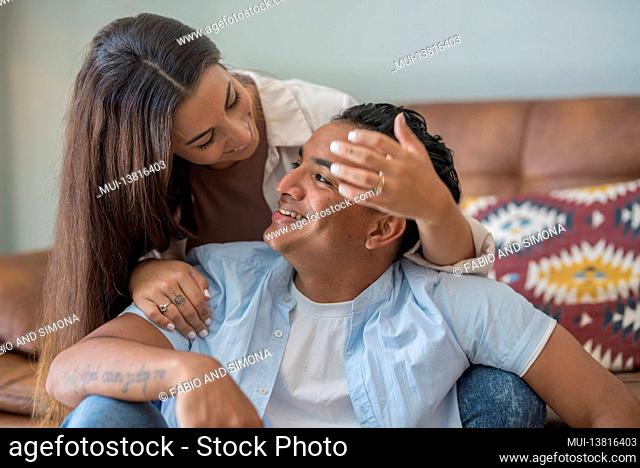 Young interracial black caucasian couple in love at home sitting on the sofa smile and look each other together in relationship - millennial indoor lifestyle...
