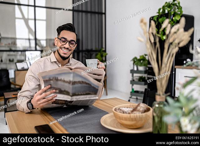 Morning news. Cheerful young bearded man in glasses sitting with coffee reading interested newspaper sitting at table in morning