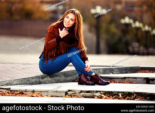 Happy young fashion woman in leather jacket sitting on city street