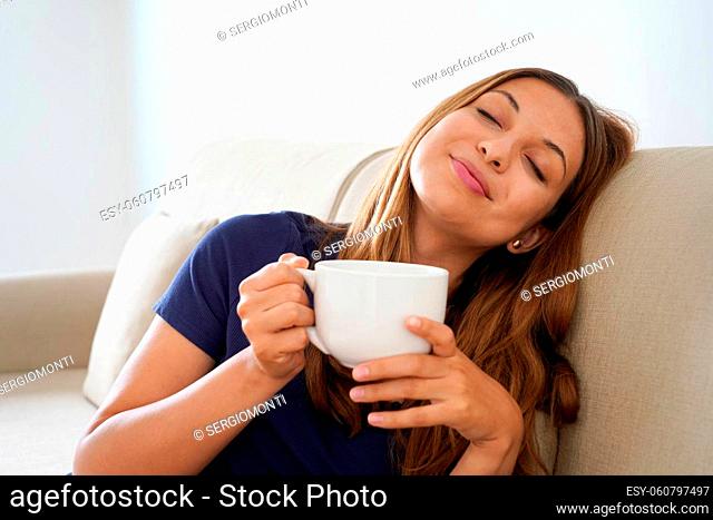 Dreamy smiling positive girl eyes closed holds cup with warm beverage imagining sitting on sofa