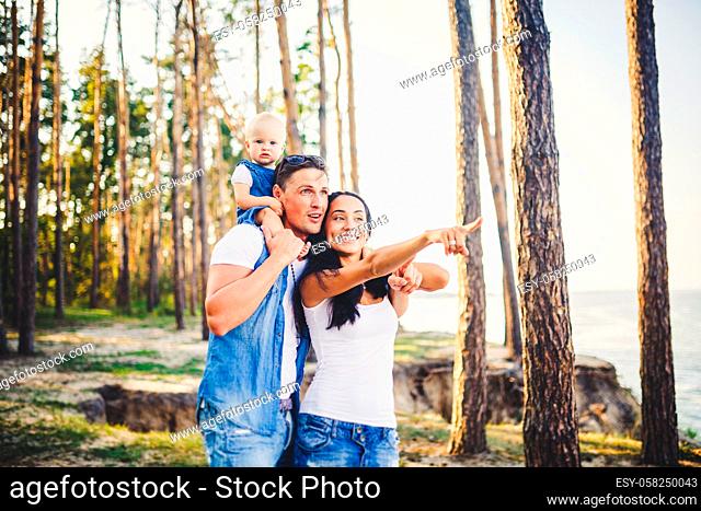 happy young family of three people resting in a park outside the city. daughter sits at daddy on shoulders, and parents show direction forward by a hand for...