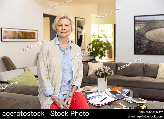 Portrait of senior Caucasian female designer working from home, sitting on back of couch holding glasses with fabric swatches and notes laid out around her