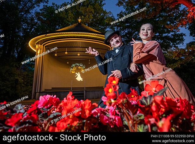 08 October 2021, Saxony, Bad Elster: Actors dressed as Charlie Chaplin and Anna May Wong pose in front of the illuminated bandstand at the Royal Spa House in...