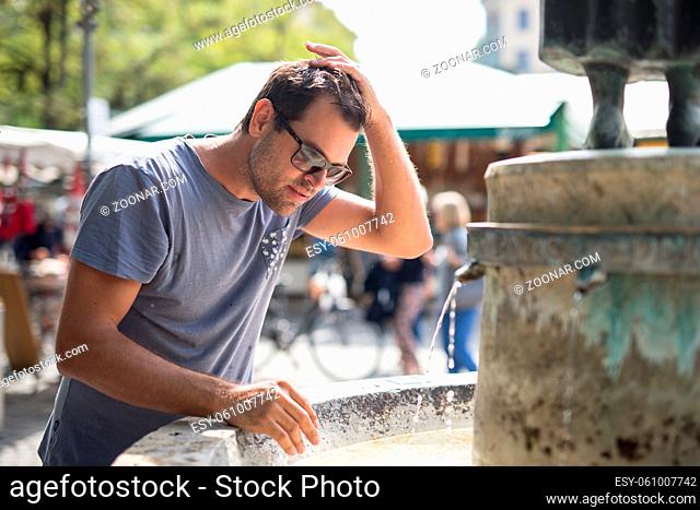 Young casual cucasian man refreshing himself with water from public city fountain on a hot summer day