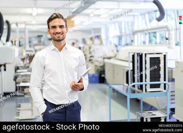 Smiling confident young businessman holding digital tablet while standing with hands in pockets at illuminated factory