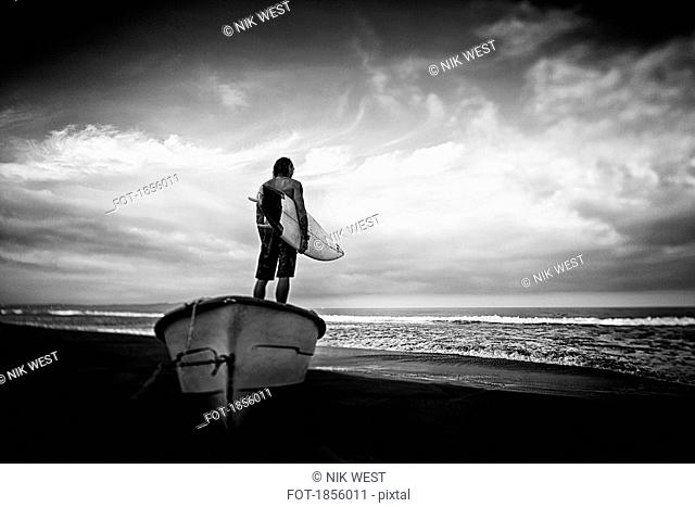 Male surfer with surfboard standing on beached boat, Higuera Blanca, Nayarit, Mexico