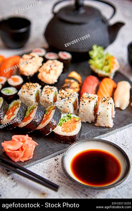Assortment of different kinds of sushi rolls placed on black stone board. Traditional asian iron tea pot on side. Top angle view