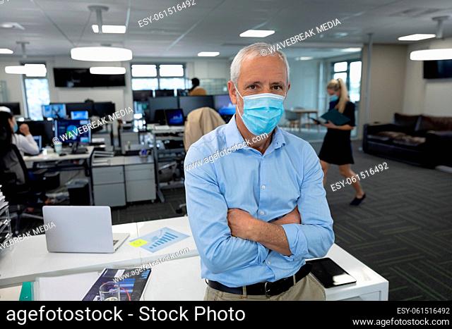 Portrait of senior businessman wearing face mask with arms crossed at modern office