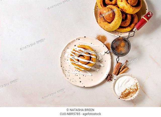 Homemade pumpkin cinnamon bun rolls sweet autumn baked dessert with cream cheese sauce in spotted ceramic plates with cinnamon sticks and vintage sieve over...