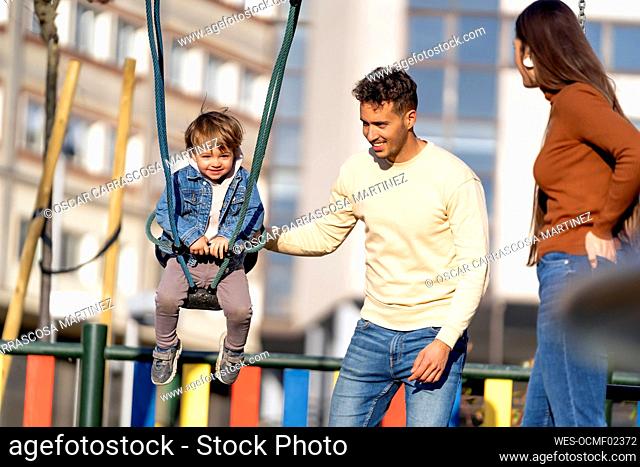 Smiling man pushing son on swing standing by woman in playground