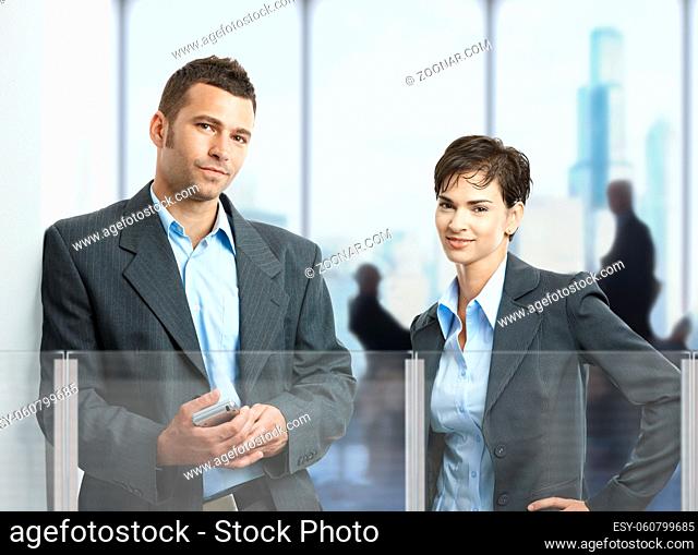 Two young businesspeople standing in corporate office lobby, looking at camera, smiling