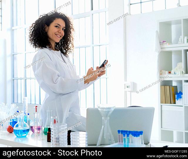 Smiling female scientist with smart phone while leaning on table at hospital