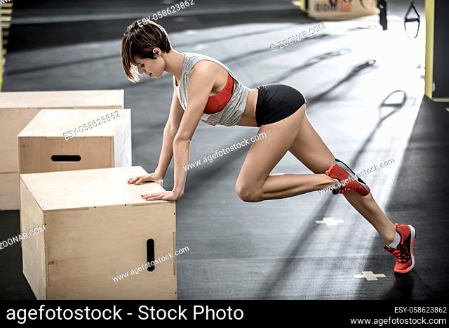 Beautiful girl leans on the wooden box with her hands and holds the left leg bent in the knee in the gym. She wears a black shorts, red top and sneakers