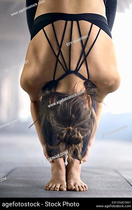Flexible girl in a black sportswear engaged in yoga on the windows background in a loft style hall. She stands on the mat and does a forward tilt and holds...