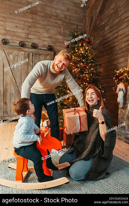 Theme family holiday New Year and Christmas. Young caucasian family mom dad son on wooden floor near fireplace christmas tree on Christmas evening