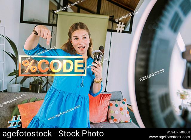 Female teenage influencer with make-up brushes giving promo code on beauty products while vlogging at home