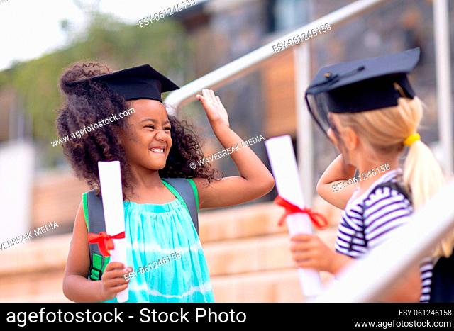 Cheerful multiracial elementary schoolgirls with mortarboards and degree doing high-five on steps