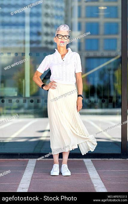 Senior woman standing with hand on hip against glass window in city