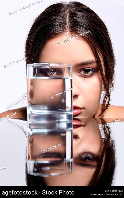 Girl hides her face behind a glass with water. Beauty portrait of young woman at the mirror table. Female on gray background