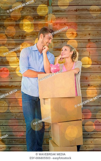 Composite image of attractive young couple leaning on boxes with piggy bank