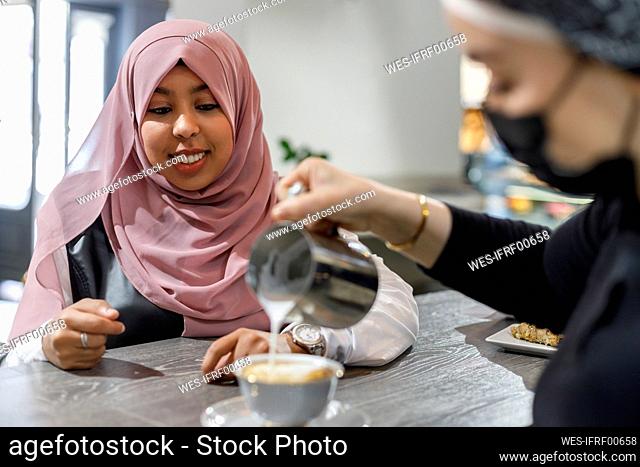 Smiling woman waiting while waitress pouring milk in cup at coffee shop