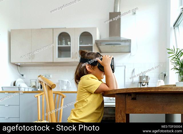 Girl wearing VR glasses sitting at table in kitchen