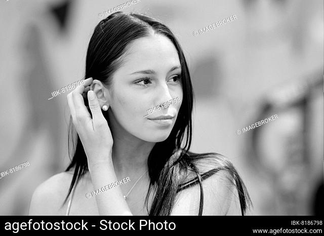 Black and white portrait of a very young pretty woman with a blurred background. With her right hand she guides her long hair behind her ear