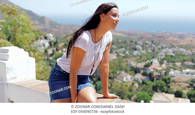 Pretty brunette in casual outfit looking excited while posing on viewpoint and exploring coastal city from height in summertime