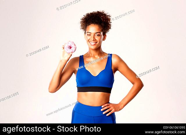 Cheerful fit african-american fitness woman in blue uniform, winking and showing donut, standing against white background