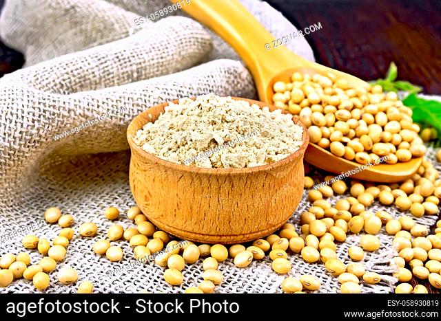 Soy flour in the bowl, soybeans in a spoon on a napkin of burlap, green soya leaf against a dark wooden board