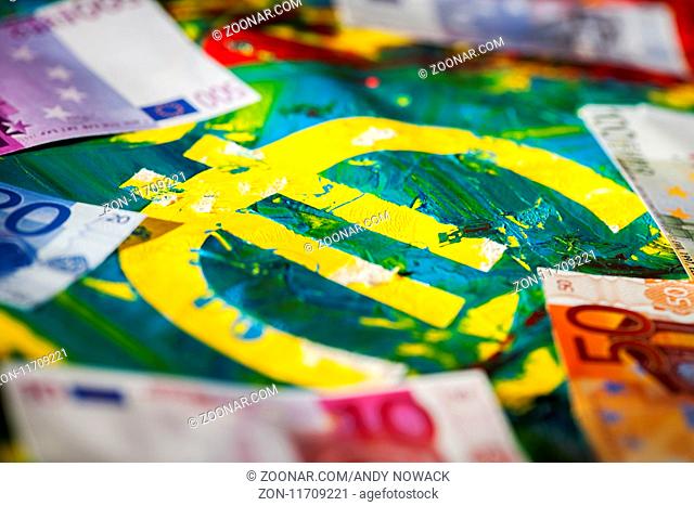 Euro-Zeichen in bunter pastös vermischter Acryl-Farbe mit aufgelegten Euro-Banknoten in Flachwinkel-Ansicht. Low angle view of a euro sign in colorful pasty...
