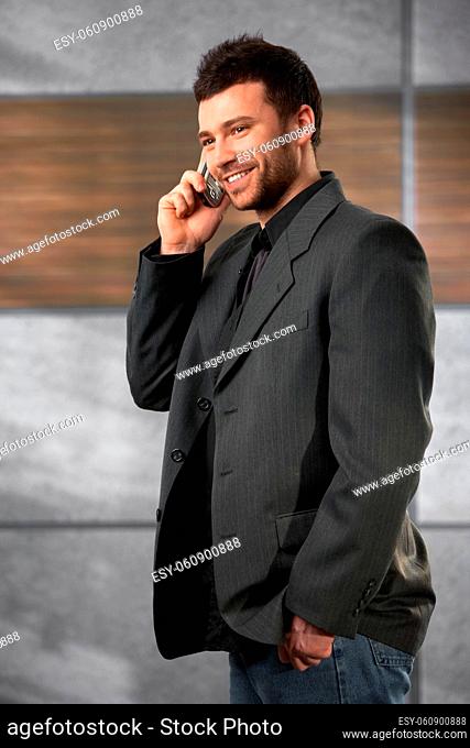 Trendy young businessman talking on mobile phone in office lobby, smiling