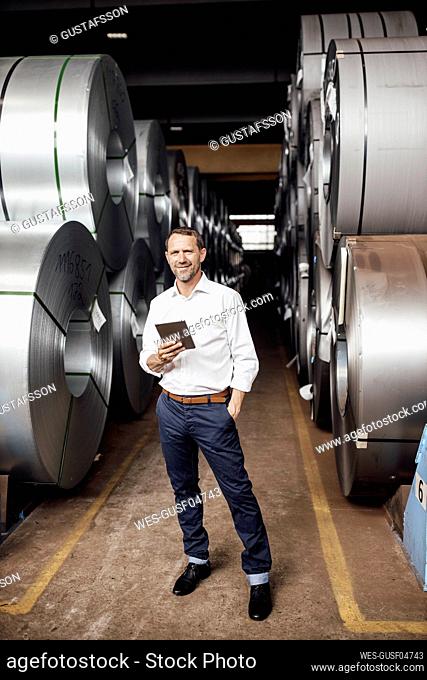 Smiling businessman with hand in pocket holding digital tablet while standing in industry