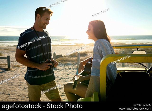 Caucasian couple on beach, man holding camera smiling, woman sitting on beach buggy playing guitar