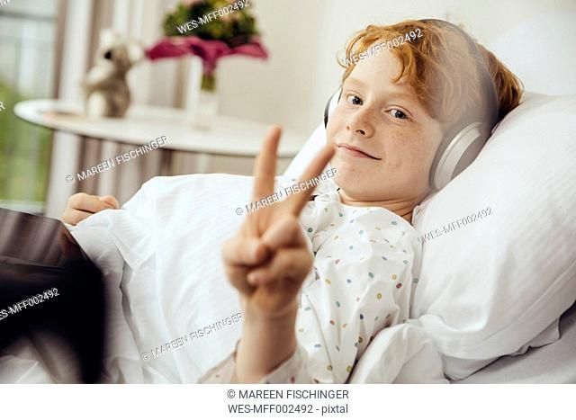 Sick boy lying in hospital making victory sign, wearing head phones