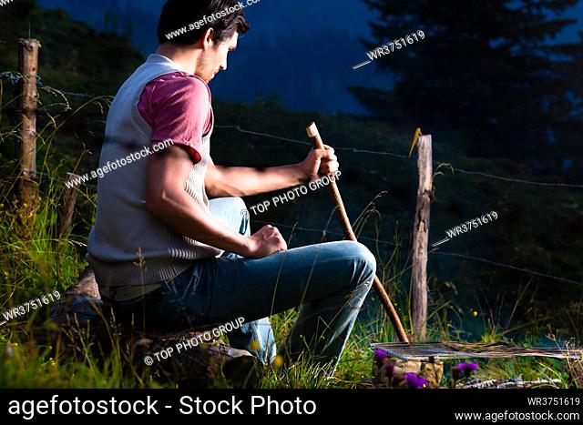 Tyrol - Young man sitting on alpine meadow of a mountain on Campfire in the Bavarian Alps and enjoys the romantic evening sunset of the panorama in leisure time...