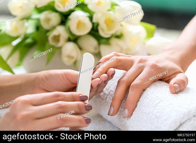 Close-up of the hands of a qualified manicurist filing the nails of a young woman with a white buffer in a trendy nail salon