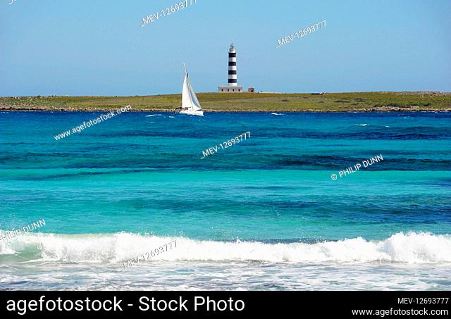 A yacht with reefed sails heels over as it beats to windward in turquoise sea towards the lighthouse on Isle Del Aire, Menorca, Spain